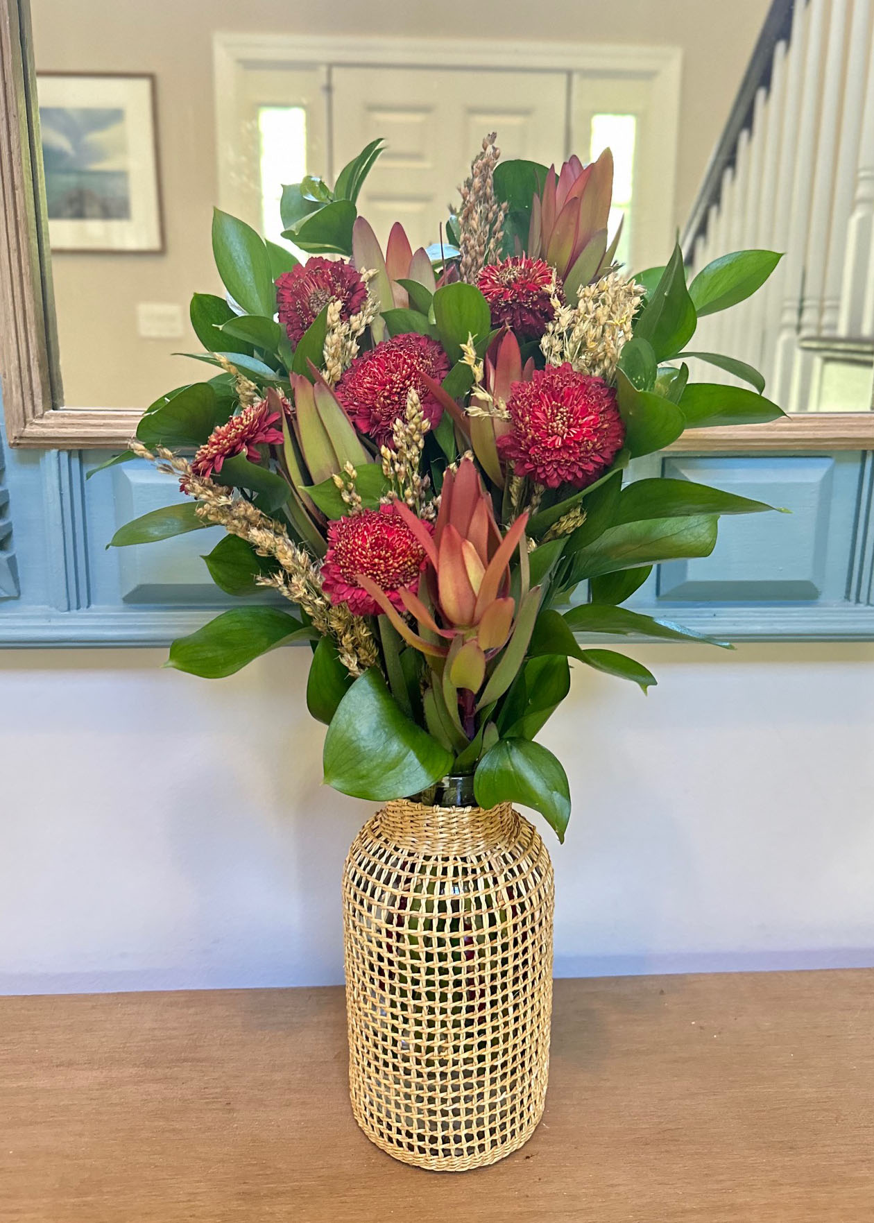 Protea, Red Mums, Millet, Israeli Ruscus, Shiney greenery, floral arrangement/arranging, floral accents, flower, autumn/fall, home decor, floral design, wicker vase, autumn/fall vases, Fall Flower arrangement in front of blue mirror